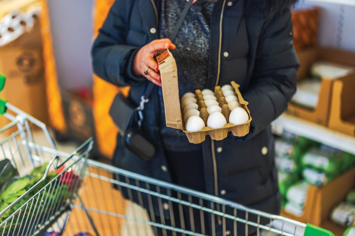 Woman in black winter coat holding carton of white eggs while shopping in grocery store aisle. Sweden.