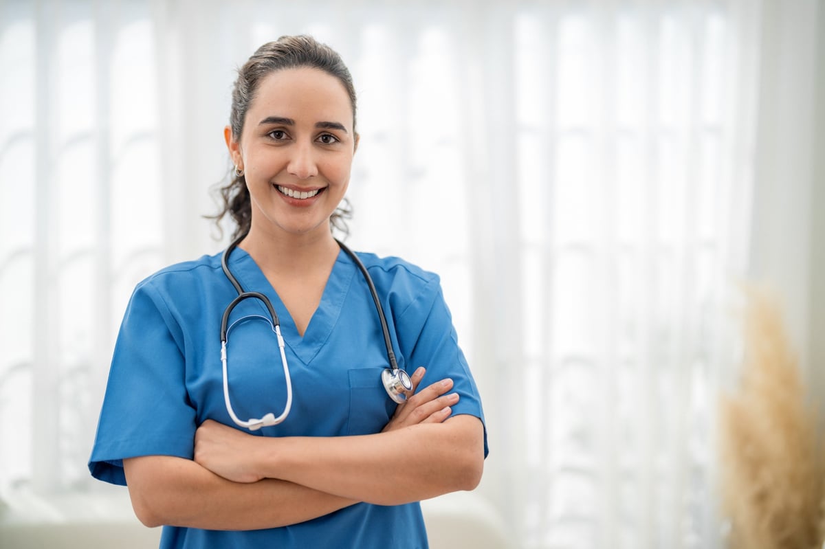 A healthcare professional stands with crossed arms, wearing blue scrubs and a stethoscope around her neck. She smiles warmly in a well-lit, contemporary office space.