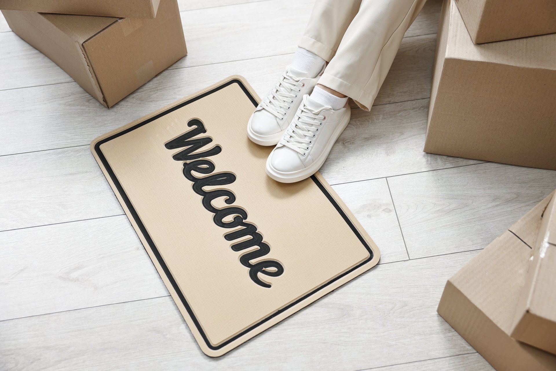 Woman sitting near cardboard boxes and doormat with Welcome in her new home