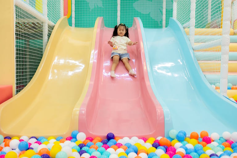 A joyful young Asian girl wearing a cream t-shirt and denim shorts enjoys playing on a pink slide, landing into a large pool of vibrant colorful plastic balls at a modern indoor soft play center for kids.