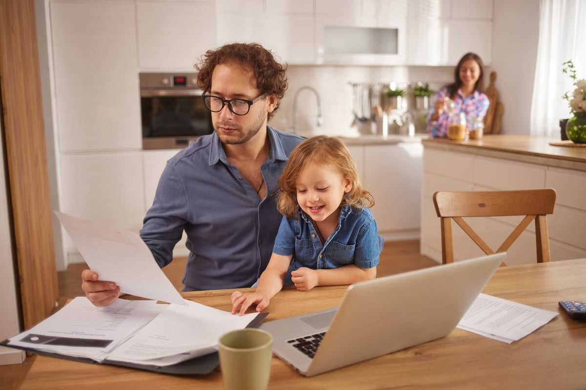 Father multitasking, working with documents while his child is learning and mother is in the background