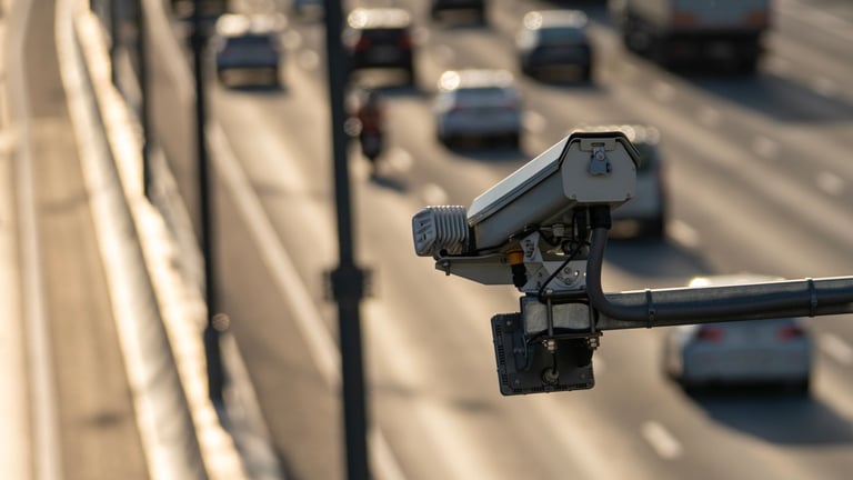 Traffic camera mounted on a highway pole. Cars flow on the multi-lane road. Sunlight reflects off the camera surface. Surveillance equipment monitors road traffic. Ideal for traffic management content