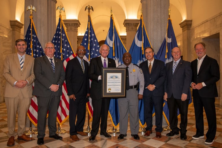 Group of nine men in formal business attire posing with a framed certificate beneath American and blue flags in a columned government building.