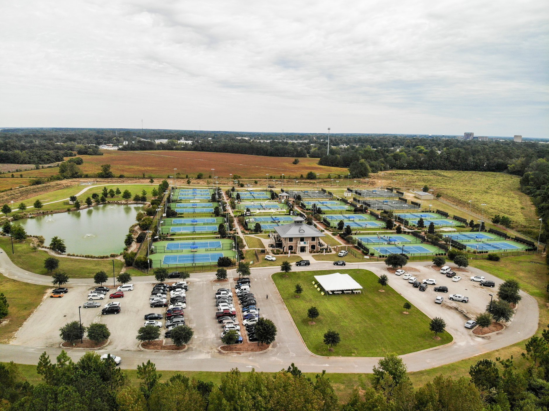 Aerial view of a tennis facility with multiple courts, parking lot, and surrounding green fields