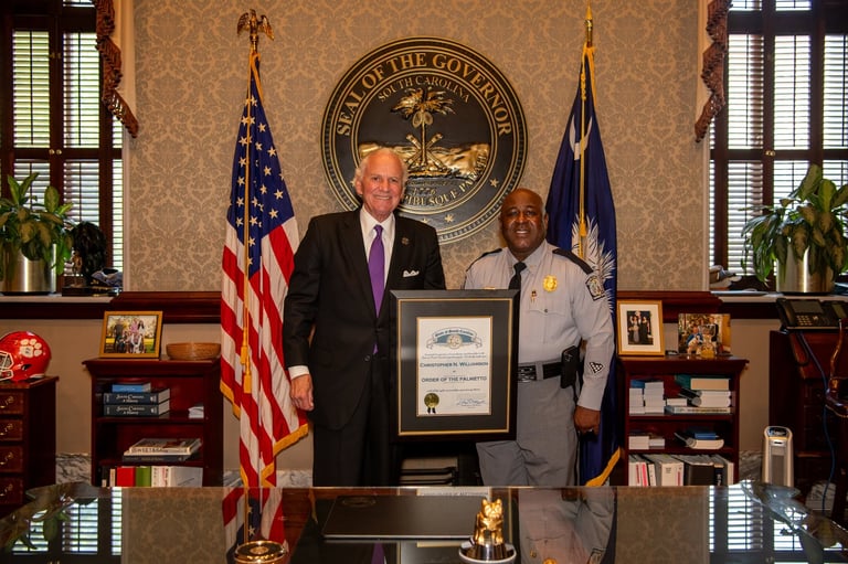Two men in formal attire hold a framed certificate in an official government office with flags and state seal.