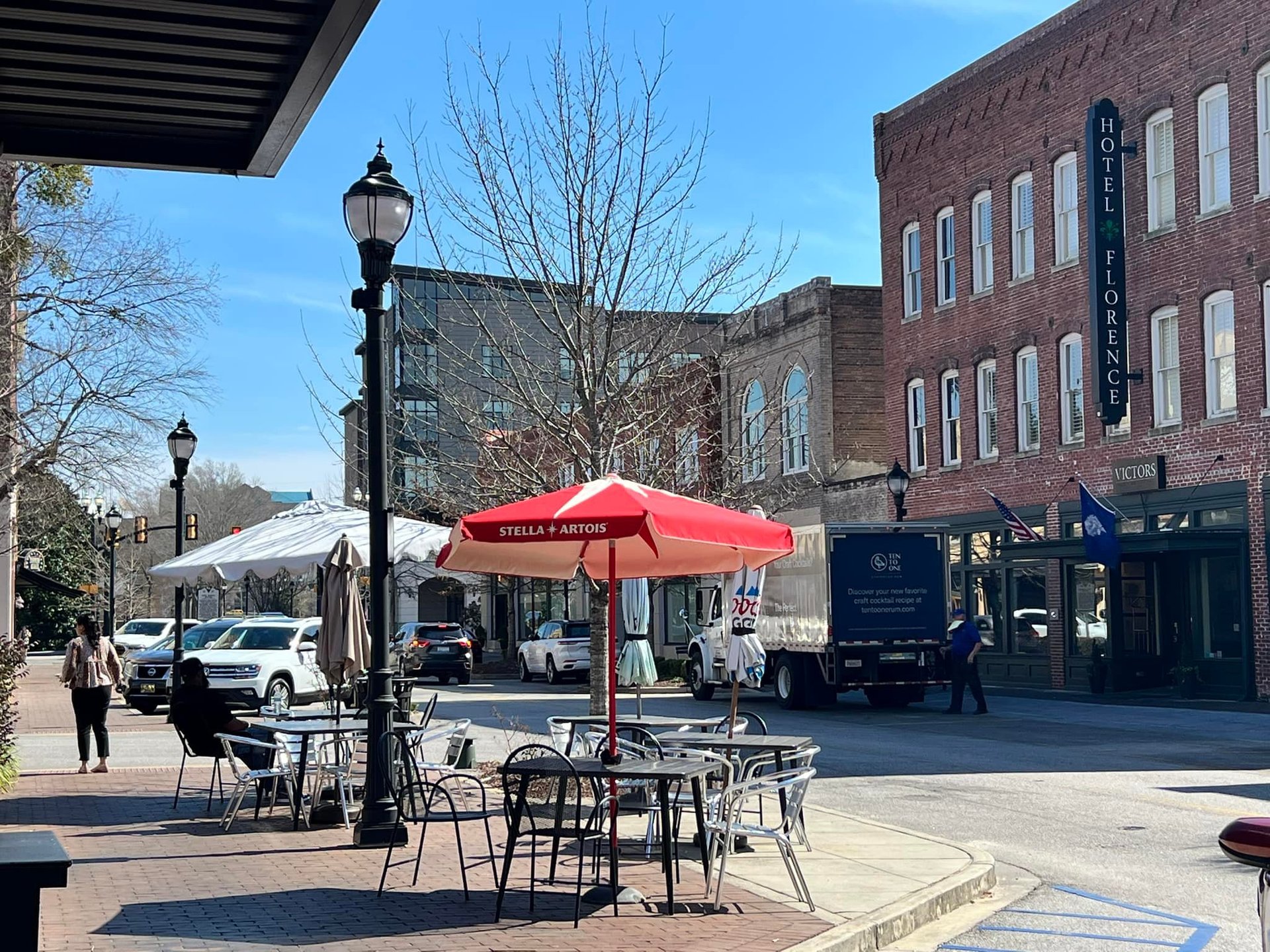 Outdoor seating area under red umbrella on brick street with historic buildings and market tent in background