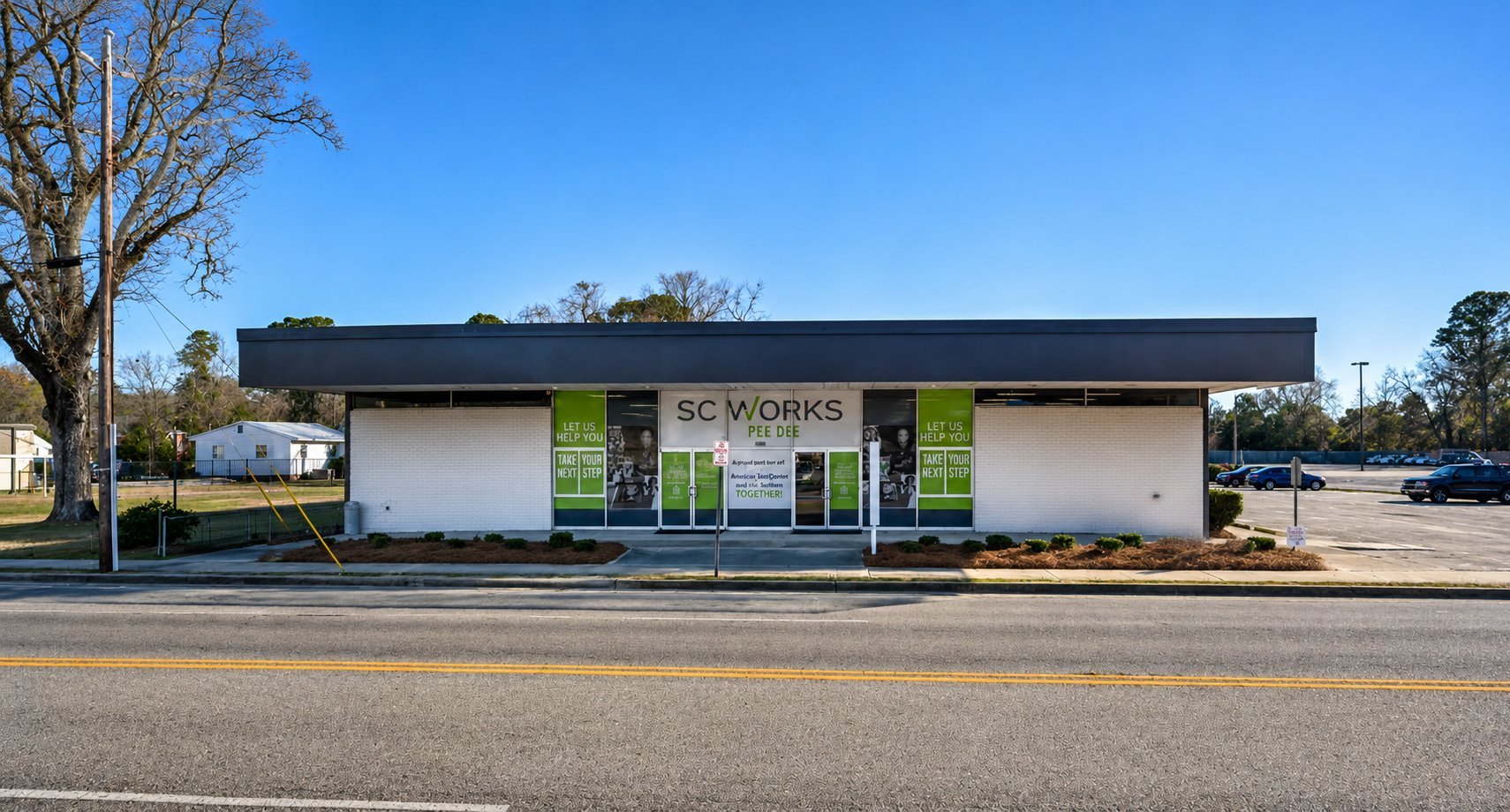 Modern single-story commercial building with white walls, black roof, and green accents on a roadside lot with clear blue sky