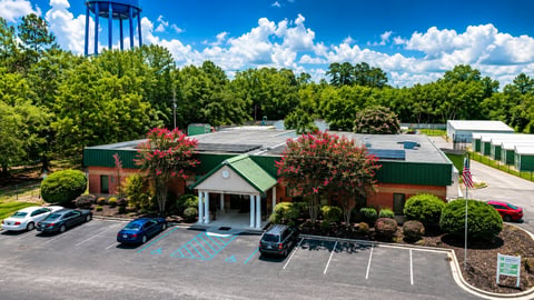 Aerial view of a brick commercial building with green roof, entrance portico, parking lot with vehicles, and surrounding landscaping on a sunny day