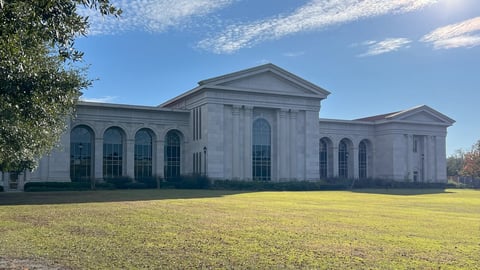 A large white neoclassical building with columns and multiple arched windows sits on a wide green lawn under a blue sky.
